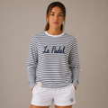 A woman modeling a navy and white horizontally striped long-sleeve t-shirt with 'Le Padel' embroidered across the chest, paired with white shorts, against a solid beige background.